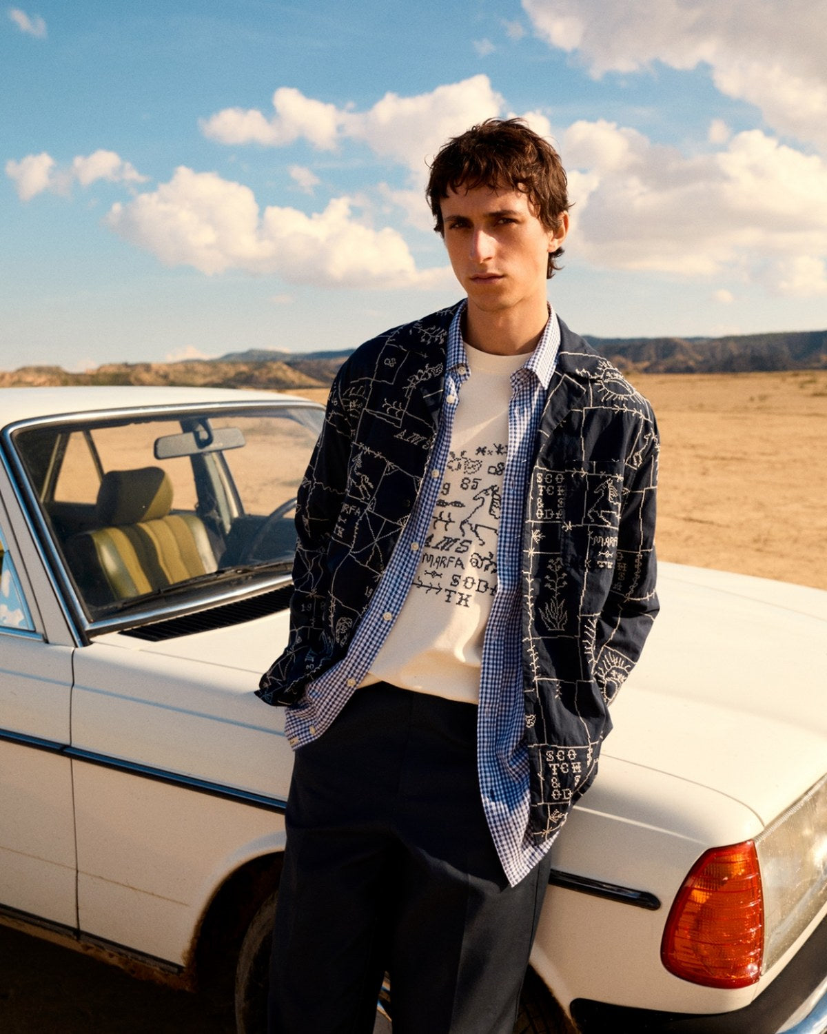A man in a Scotch & Soda embroidered navy overshirt leaning against a vintage sedan in the desert.